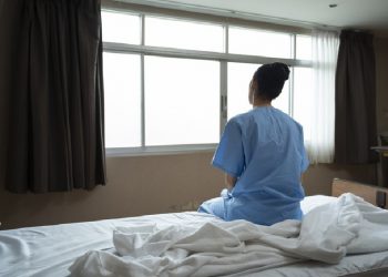 Mulher sentada em cama de hospital olhando pela janela